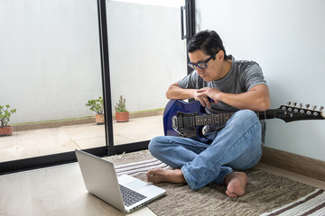 Man sitting on a carpet with an electric guitar tentatively looks at his computer