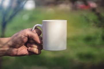 Man holding a ceramic white cup of coffee or tea outdoors. Man's hand holding a porcelain mug against green grass background. Close up, mockup image, copy space for text.