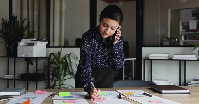 Young busy indian business woman working on project paperwork talking on the phone. Ethnic female manager making business call on mobile writing tasks on sticky notes working alone in office.
