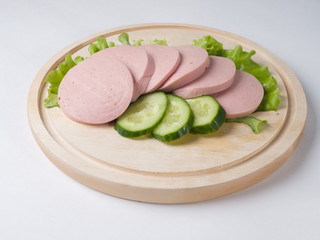 Boiled sausage, cut into slices on a board. Near lettuce leaf and fresh cucumber