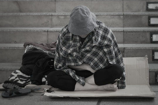 Homeless man sitting on the street in the shadow of the building. He begging for  help and money. Poverty concept. Black and white picture