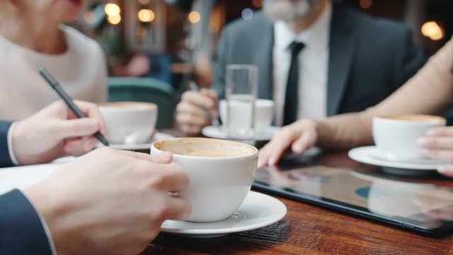 Group of business people are drinking coffee and talking at meeting in restaurant, close-up of cup and hand holding it with wooden table in background.