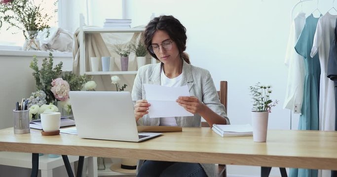 Smiling female fashion designer opening envelope reading mail letter sitting at desk in modern cozy studio. Happy business woman clothing store owner entrepreneur checking correspondence concept.