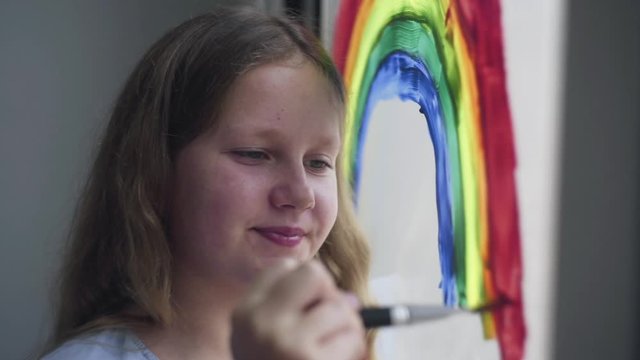 Girl painting rainbow during Covid-19 quarantine at home. Girl near window. Stay at home Social media campaign for coronavirus prevention, let's all be well, hope during coronavirus pandemic concept
