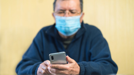 grandpa with cell phone sitting using his phone. man with smartphone sitting indoors at home due to quarantine. Mister looking at his phone. Lonely concept. Quarantine, Coronavirus, face mask