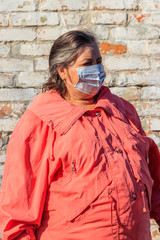 Senior woman stands in a medical mask on her face, which protects against coronavirus and other viruses and diseases on a background of old brick wall. Coronavirus pandemic