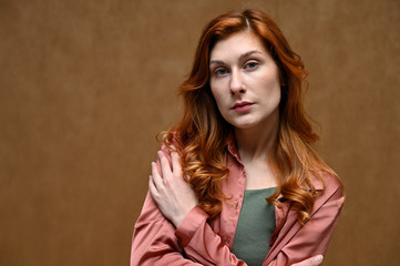 Photo portrait on a beige background of a pretty young woman in a pink shirt with long beautiful red hair. The model shows pensive emotions.