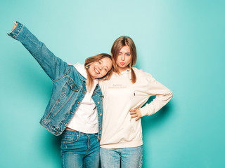 Two young beautiful brunette smiling hipster girls in trendy summer hoodie and jeans jacket clothes. Sexy carefree women posing near blue wall. Trendy and positive models having fun