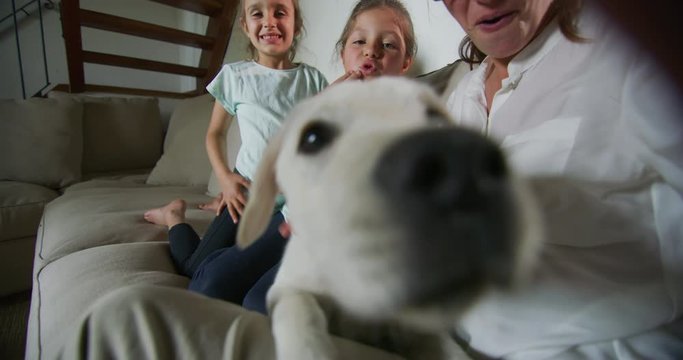 Authentic shot of happy mother with her kids and Labrador puppy are making a selfie or video call in living room. Concept of technology, new generation,family, connection, parenthood, authenticity