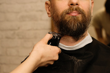 Bearded man with unshaven beard, brutal hipster guy with moustache, getting stylish beard shaving with electric razor by barber at barbershop. Details of trimming. Close up