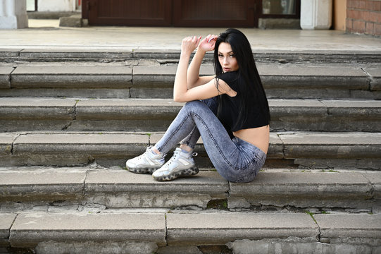 Full-length photo in spring weather outdoors, caucasian brunette girl with a slim figure in jeans and a black T-shirt. Fashionable model sits on the stairs of the building.