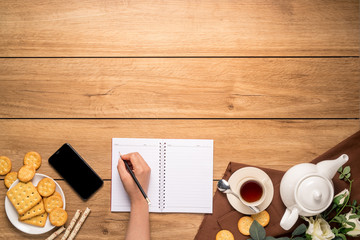 Afternoon tea set with bread, crackers and mobile phones, and a woman's hand is taking notes on the side on the wooden table, with copy space, top view.