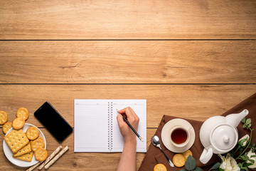 Afternoon tea set with bread, crackers and mobile phones, and a woman's hand is taking notes on the side on the wooden table, with copy space, top view.