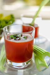 Two glasses of tomato juice with parsley and celery decorations, stand near the window, morning sunlight shines, shallow depth of field, selective focus. Natural drinks concept.