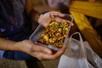 Blurry image a woman wearing a blue dress, holding a plastic box, wearing an oyster salad bought from a restaurant on the pedestrian street. There is a white garbage bag next to it.