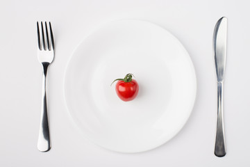 Eating a low calorie diet concept. Top above overhead flat-lay view photo of a plate with a single tomato with fork and knife aside isolated on white background