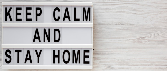 'Keep Calm and Stay Home' words on a lightbox on a white wooden background, top view. Overhead, from above, flat lay. Copy space.