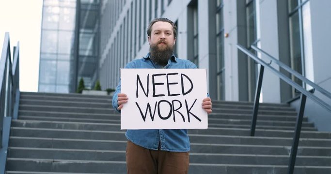 Portrait shot of Caucasian young jobless man with beard standing outdoor at big stairs and showing table with words Need Work. Male workless demonstrating board with protest against unemployment.