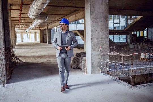 Smiling attractive supervisor in suit, with helmet on head and tablet in hands walking in building in construction process and evaluating work.