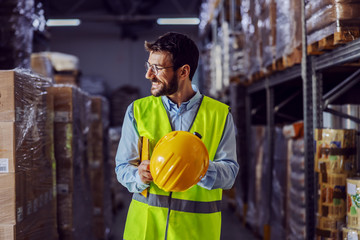 Young smiling supervisor in vest, helmet in his hands and folder under armpit going around...