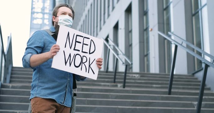 Young Caucasian male activist in medical mas standing at street of city on big stairs with poster Need Work. Hard pademic times. Unemployment protest concept.