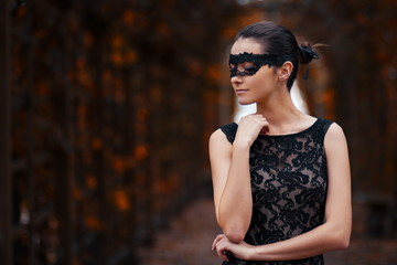 elegant girl in a black mask and a long black dress posing in the park in autumn