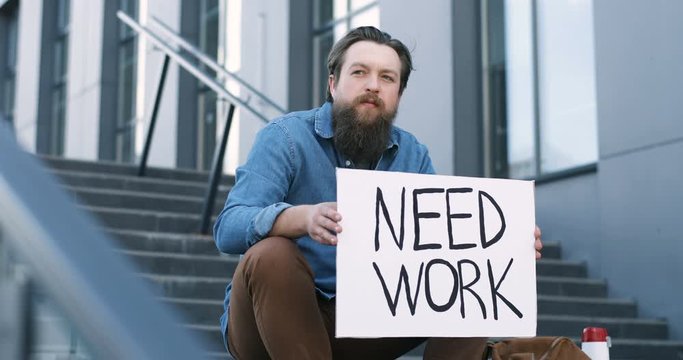 Portrait of young workless Caucasian man with beard sitting on stairs outdoors and holding carton table Need Work. Unemployment after pandemic lockdown. Workless male.
