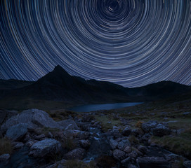 Digital composite image of star trails around Polaris with Stunning landscape image of countryside around Llyn Ogwen in Snowdonia