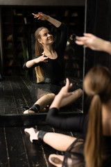 Fototapeta na wymiar Beautiful ballerina in black dress warming up before a performance at the theatre. The girl's reflection in the mirror.