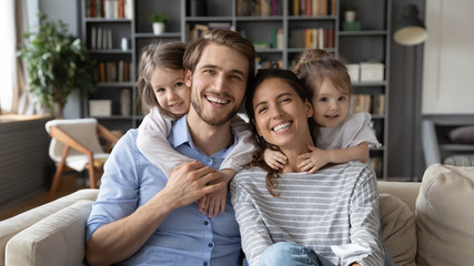 Portrait of young Caucasian family with small daughters pose relax on sofa in living room, smiling little girls kids hug embrace parents, show love and gratitude, rest on couch at home together