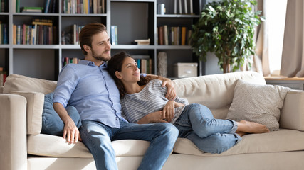 Fototapeta premium Happy young Caucasian couple relax on cozy sofa at home hugging look in distance dreaming together, dreamy millennial man and woman rest on comfortable couch embracing, enjoy weekend at home