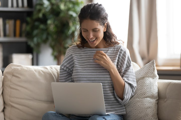Happy young woman sit on sofa in living room have fun shopping online on laptop or browsing internet, smiling female feel excited reading good unexpected news or email using computer at home