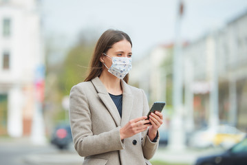 A girl with long hair in a medical face mask to avoid the spread coronavirus uses a smartphone in the street. A woman in a face mask against COVID-19 wears a coat waits in the center of the city.