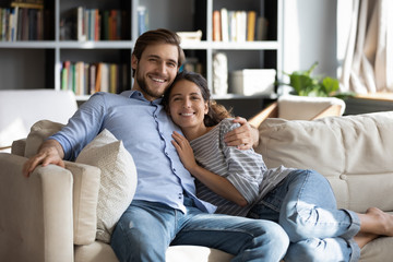 Obraz premium Portrait of smiling young husband and wife relax on cozy sofa in living room, hug and cuddle on weekend at home, happy Caucasian couple rest on couch embracing, enjoy family leisure weekend together