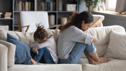 Stubborn young mom and little daughter sit on couch back to back avoid talking after family fight, offended mother and small girl child ignore each other after quarrel, generation gap concept