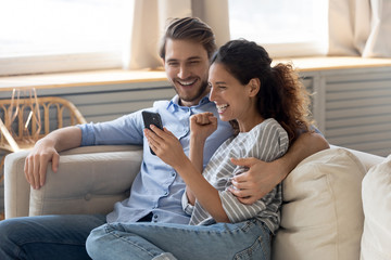 Happy young Caucasian couple relax on couch feel excited reading good news on smartphone, overjoyed...