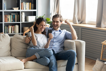 Overjoyed young Caucasian couple sit relax on couch in living room triumph winning online lottery on cellphone, happy man and woman feel excited reading good unexpected pleasant news on smartphone
