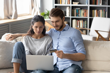 Young Caucasian couple sit relax on couch in living room shopping online on laptop paying with credit card, millennial man and woman make internet payment, buying on web using computer