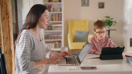 mother teleworking on laptop while son is distance learning on tablet in headphones Spbas. freelancer mom and child e-learning education in living room. concept quarantine. modern millennial family