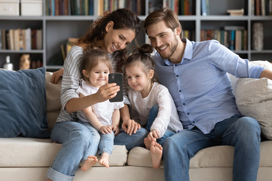 Smiling young Caucasian family with little daughters sit on couch in living room male selfie on smartphone together, happy parents with small children pose for self-portrait picture on cellphone