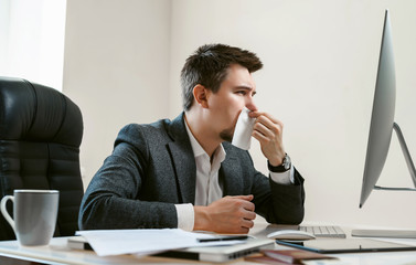 A young businessman or office worker sneezes, coughs, blows his nose at the workplace. Overworked worker got colds and flu. High temperature with ARVI or SARS. Epidemic of influenza or coronavirus.