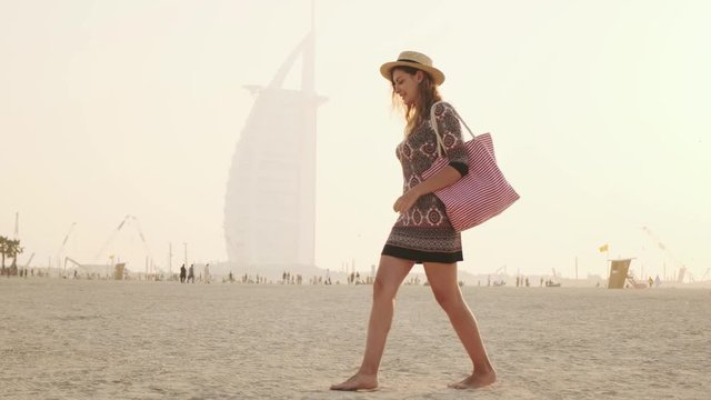 Young beautiful cute woman tourist walking sunset along shore Persian Gulf, sandy plage, coastline Dubai Beach. Girls blonde long hair. Backdrop summer sky, foggy image modern skyscraper. UAE 2020 4k