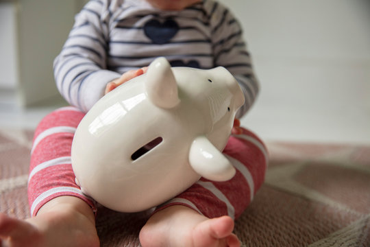 Cost of childcare concept. Young baby holds onto a white piggy bank