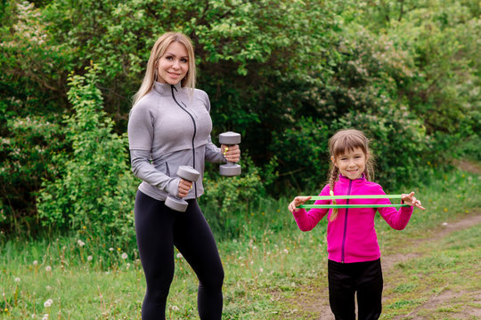 Mom and daughter 5 years old in sportswear are training in the park with dumbbells and expanders