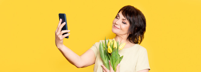 Beautiful old woman with professional smokey make-up and hairstyle holding tulip flowers on the yellow background. Taking videocall by the smartphone.