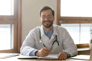 Portrait of smiling young Caucasian male doctor sit at desk filling medical journal in hospital, happy man GP or physician write make notes or patient anamnesis in card in clinic, healthcare concept - obrazy, fototapety, plakaty