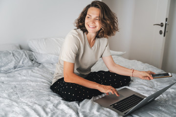 Pretty young curly woman spends time with a smartphone and a laptop on the bed in the bedroom, remote work education, dates