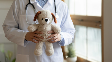 Close up of male child doctor in white medical uniform hold plush teddy bear, man pediatrician stand with fluffy toy, show care and comfort in hospital or private clinic, children healthcare concept