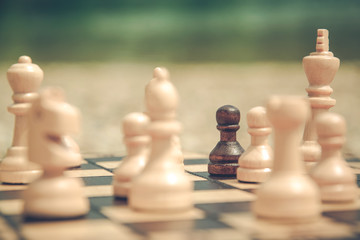 Pawn chess piece standing on a chessboard. Surrounded by white figures. Racism concept. Playing chess outdoors. 
