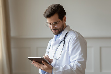 Male Caucasian doctor in white medical uniform fill patient medical history or anamnesis on tablet in hospital, man GP or physician consult client online on pad in modern clinic, healthcare concept